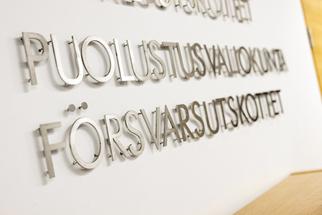 Puolustusvaliokunnan nimi suomeksi ja ruotsiksi metallinvärisin kohokirjaimin valkoista seinää vasten.