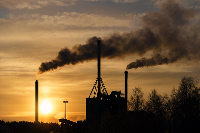 Tehtaan piippuja, joista tulee savua, taustalla aurinko matalalla taivaalla. 